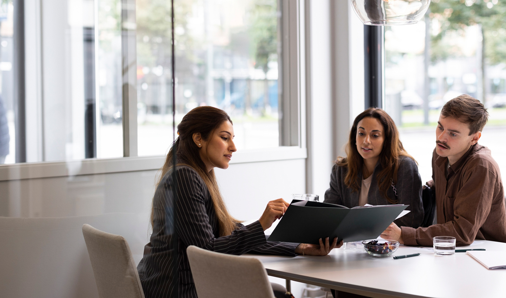 A financial advisor sits with a couple in a modern office, showing them a folder of information. The image represents the personal, one-on-one consultation and positive client relationships highlighted in our Client Stories.