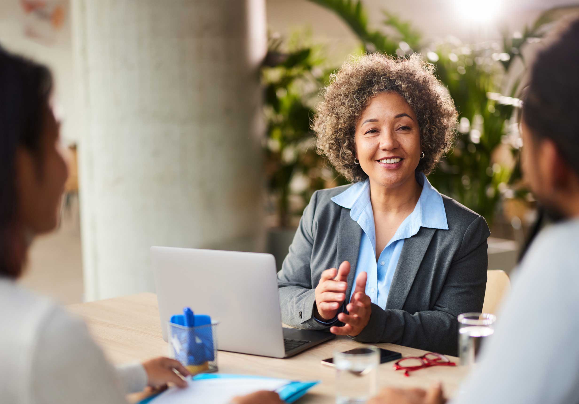 A Cliva Offshore Bank financial advisor with short curly hair is smiling and gesturing while seated at a table with her computer. She is meeting with a client to discuss their financial needs, symbolizing the expert and personalized service we offer.